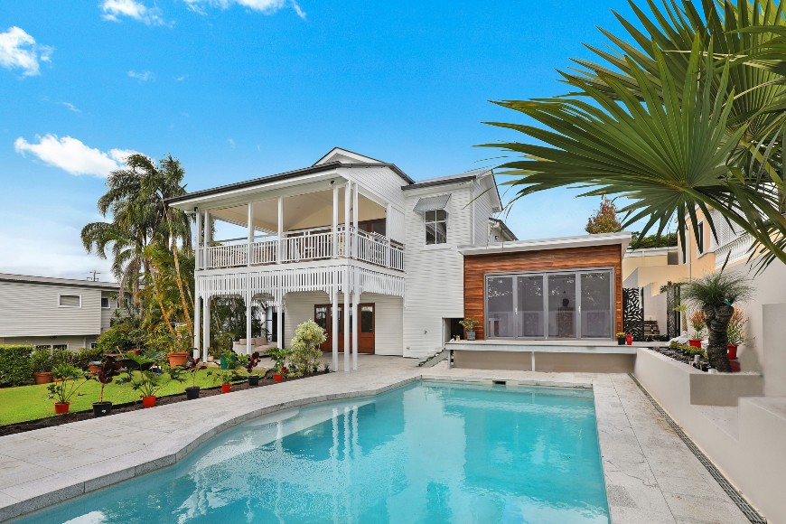 A modern two-story house with a wraparound balcony overlooks a backyard swimming pool, lush tropical plants, and a bright blue sky—perfect for those seeking fresh landscaping ideas to transform your garden.