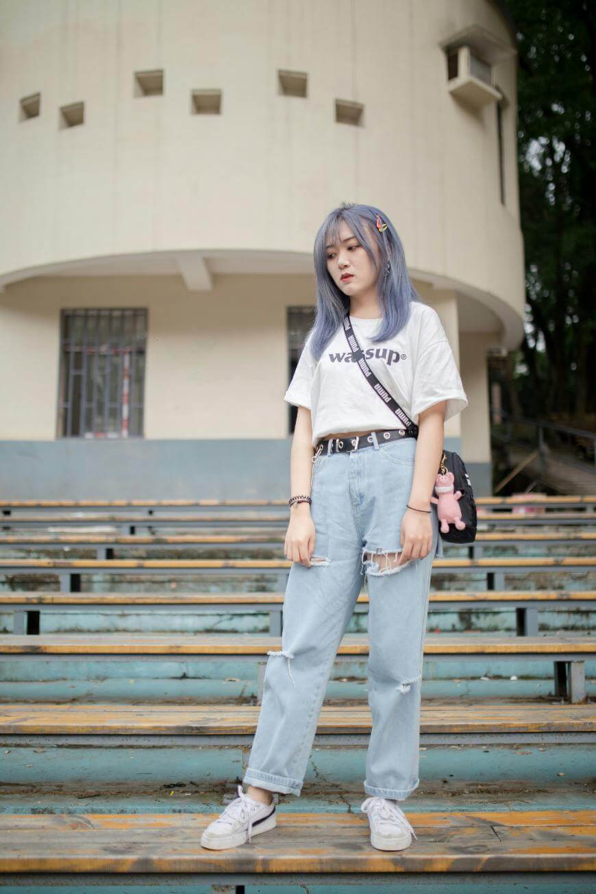 A young woman with blue hair stands outdoors on bleachers, wearing a white WASSUP t-shirt, light ripped jeans, white sneakers, and a black crossbody bag, with trees and a white building in the background.