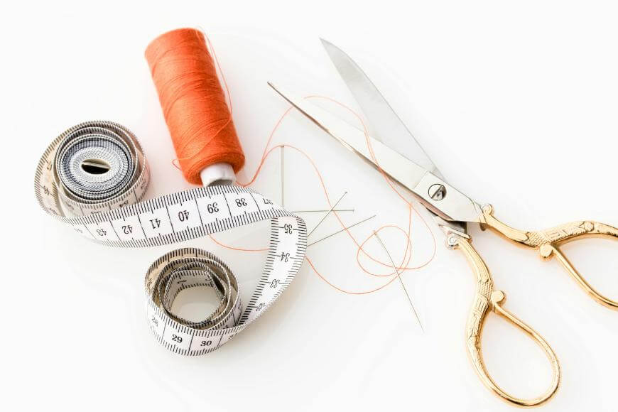 Gold-handled scissors, a spool of orange thread, a white measuring tape, and sewing needles arranged on a white surface. Some orange thread is loosely strewn around the items.