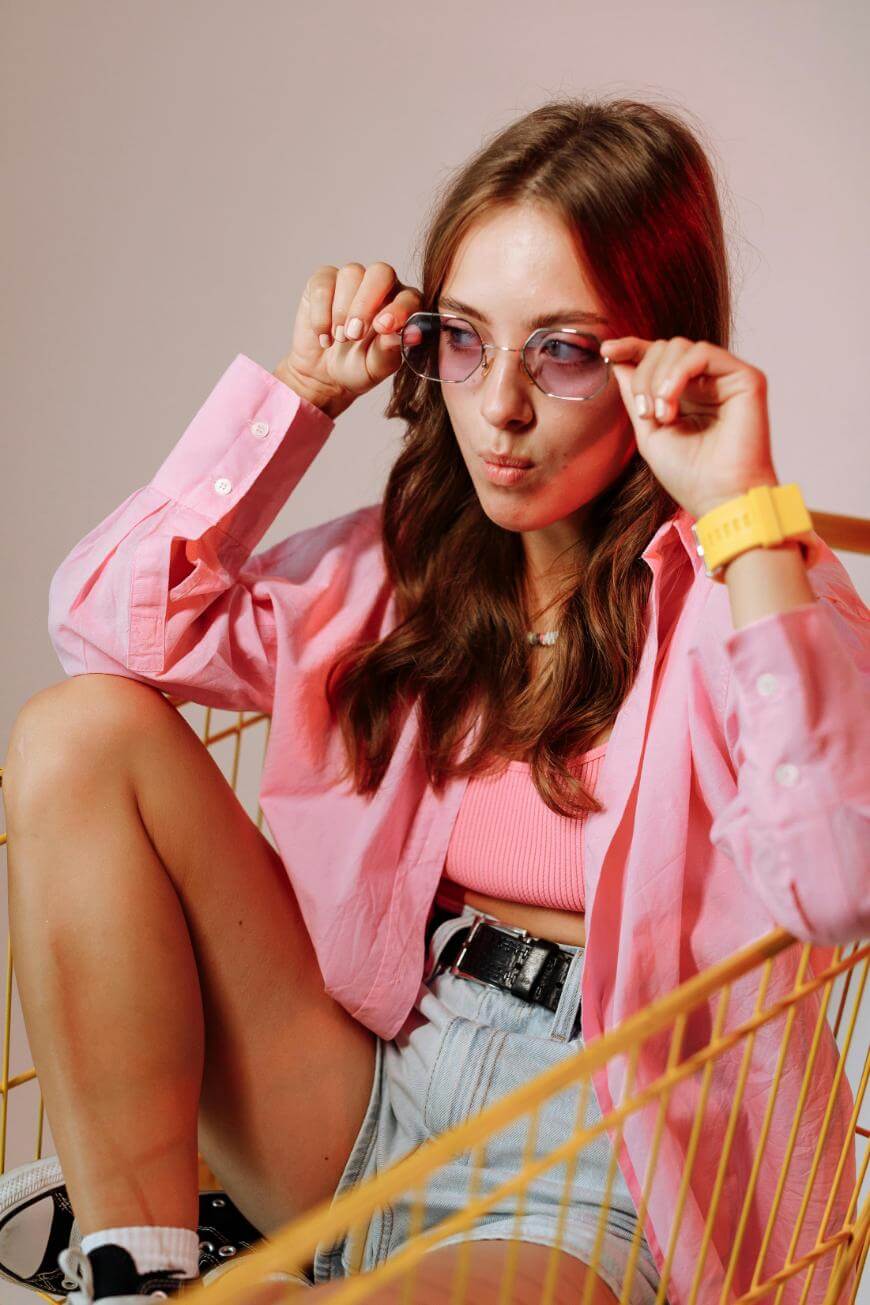 A young woman wearing a pink shirt, pink crop top, and denim shorts sits in a shopping cart. She adjusts her sunglasses and has a yellow watch on her wrist. The background is softly lit with pink tones.