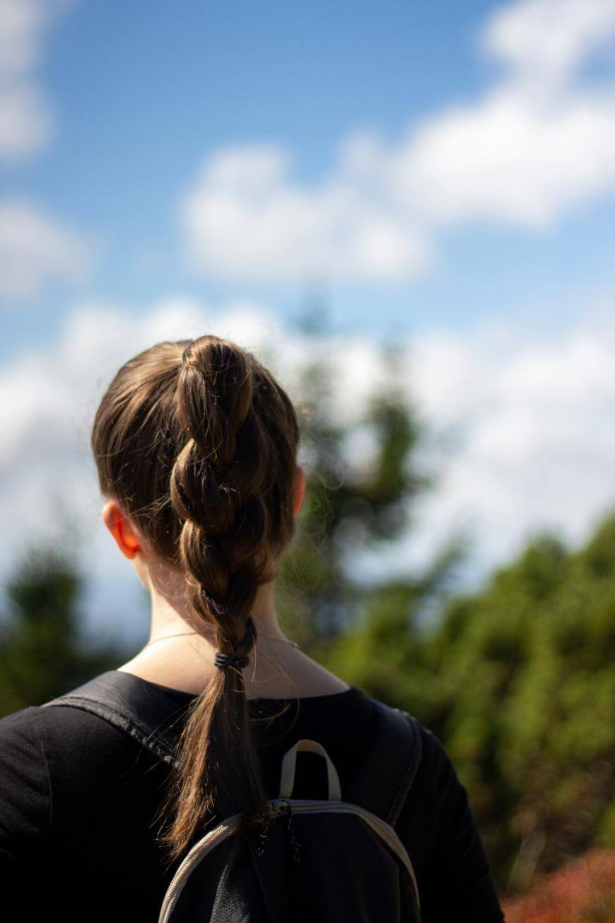A person with a braided ponytail and a black shirt stands outdoors with their back to the camera, wearing a backpack. The background is blurred, showing green foliage and a blue sky with white clouds.