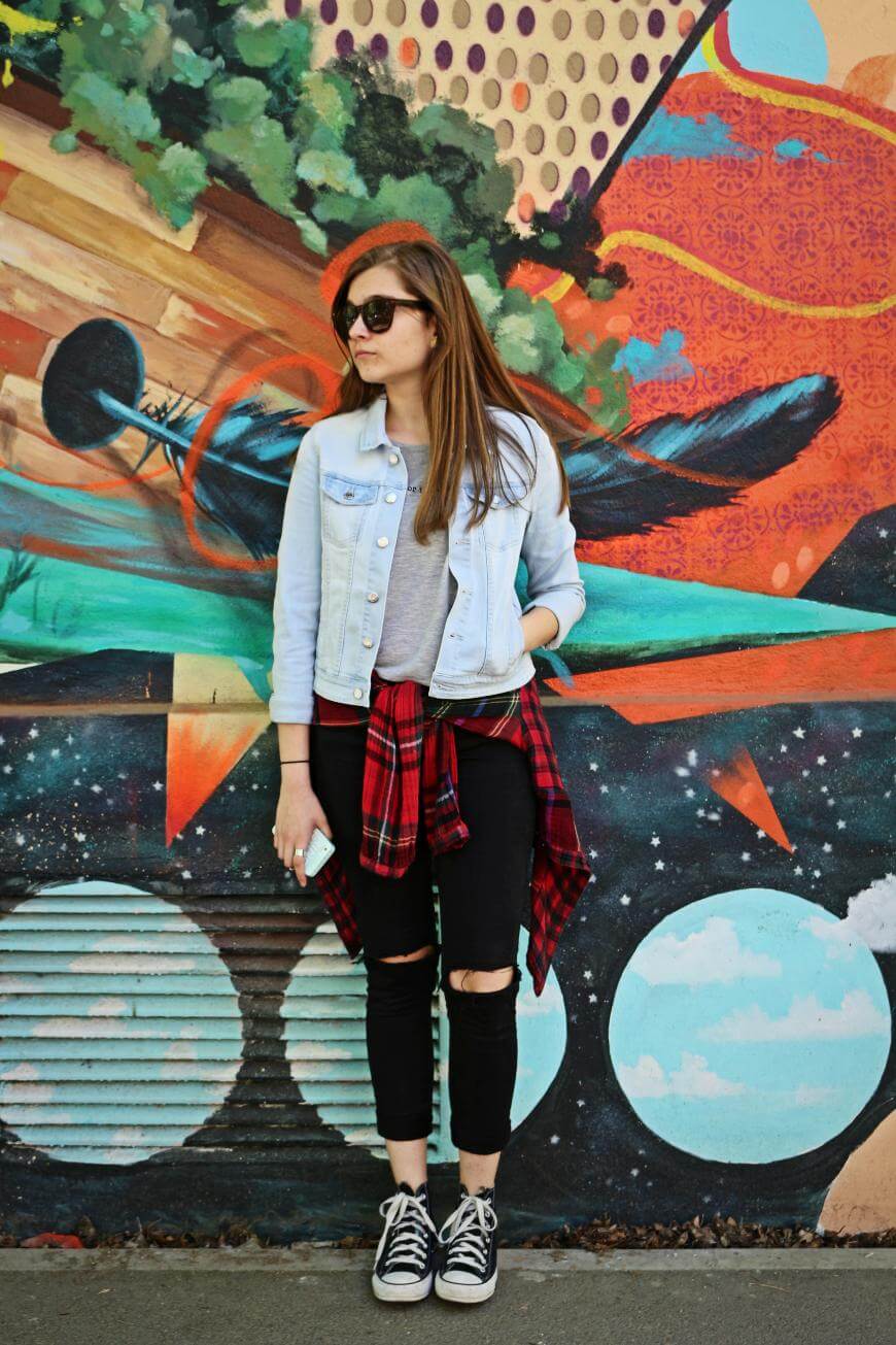 A woman in sunglasses, a denim jacket, ripped black jeans, and sneakers stands in front of a colorful graffiti wall with abstract and cosmic patterns. She has a red plaid shirt tied around her waist and holds a phone.