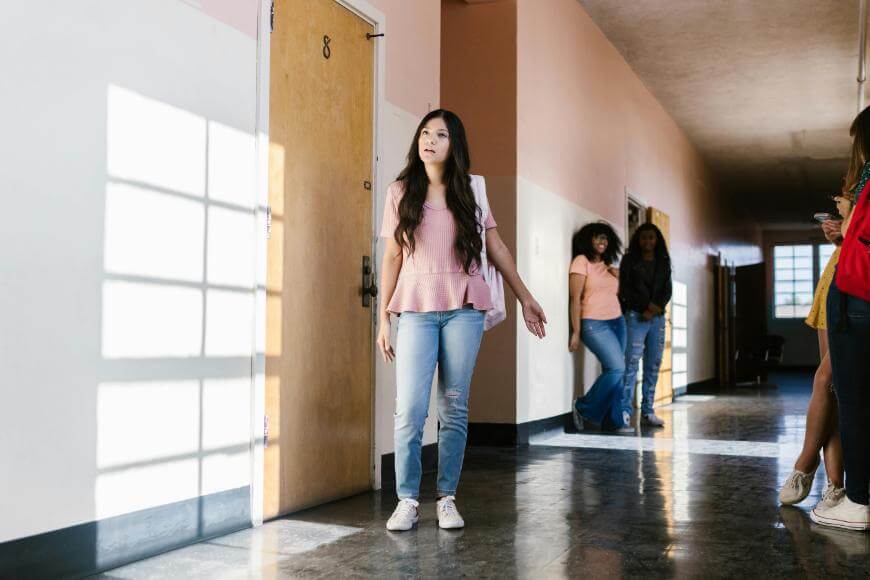 A young woman in a pink top and jeans stands alone in a bright school hallway, looking to her left. Other students are gathered and talking in the background near lockers and doors.