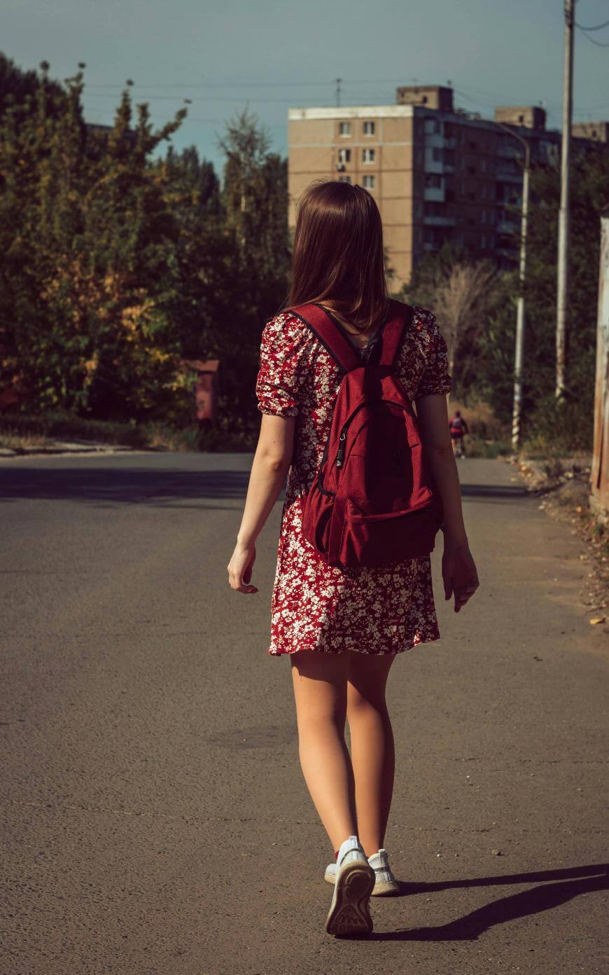 A girl with long hair, wearing a red floral dress, white sneakers, and a red backpack, is walking alone down a quiet paved street lined with trees and buildings.