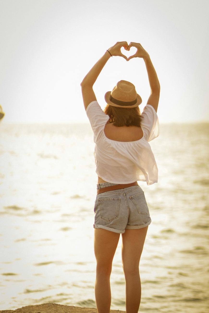 A person wearing a hat, white top, and denim shorts stands by the sea at sunset, facing the water with arms raised overhead, forming a heart shape with their hands.