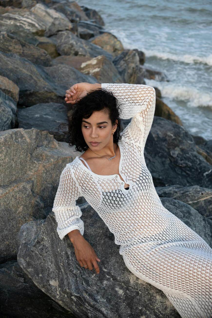 A woman in a white, crochet dress reclines on large rocks by the ocean, looking to the side with one arm resting above her head. Waves and water are visible in the background.