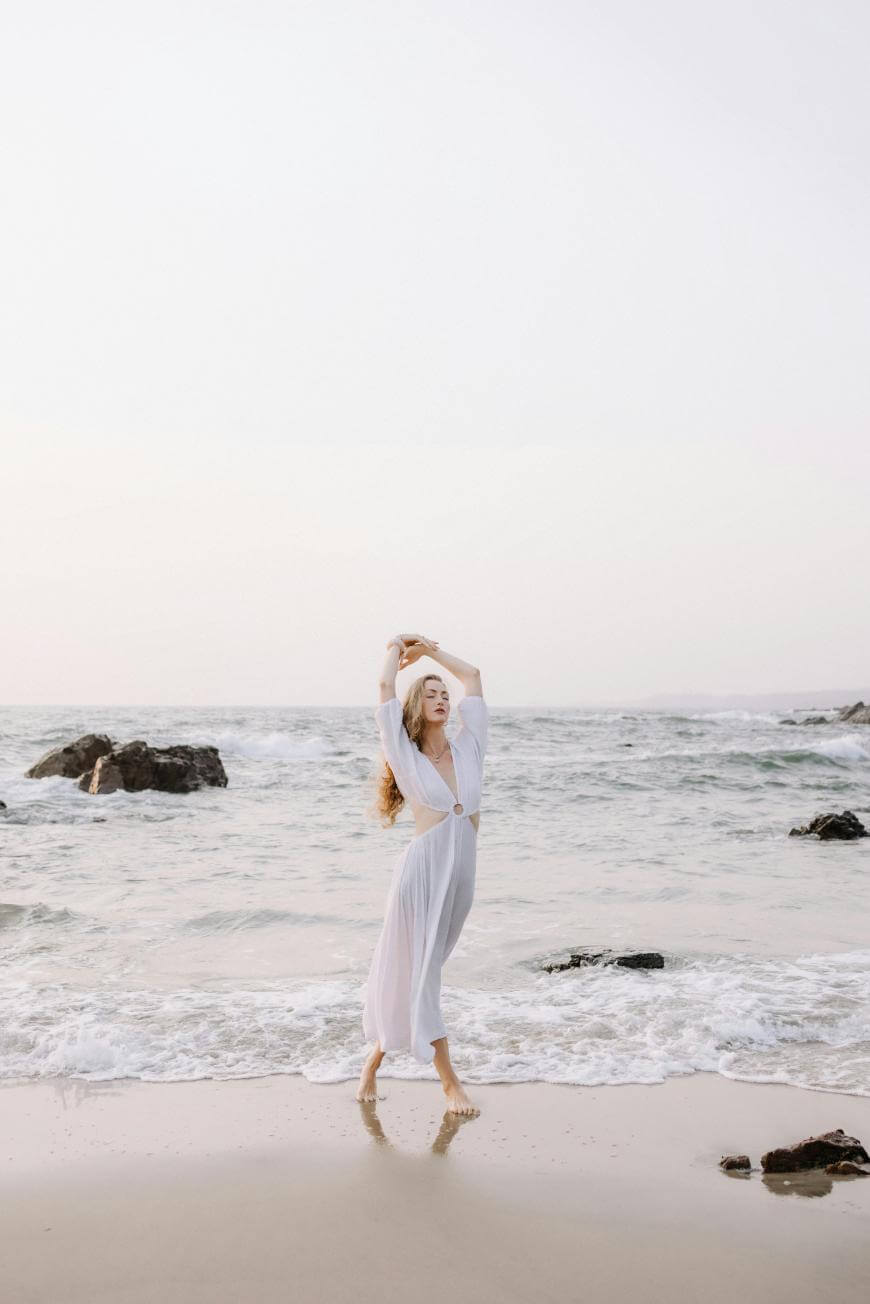 A woman in a white dress stands barefoot on the beach near the shoreline, arms raised above her head, with gentle waves and rocks in the background under a pale sky.