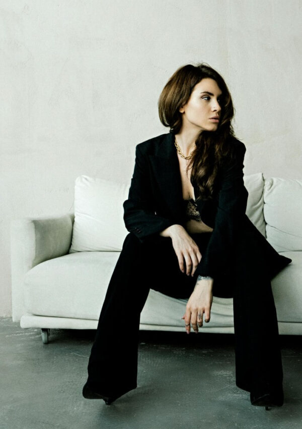 A woman with long brown hair sits on a white couch, wearing a black suit and a gold necklace. She looks to the side with a serious expression, and the background is a plain, light-colored wall.