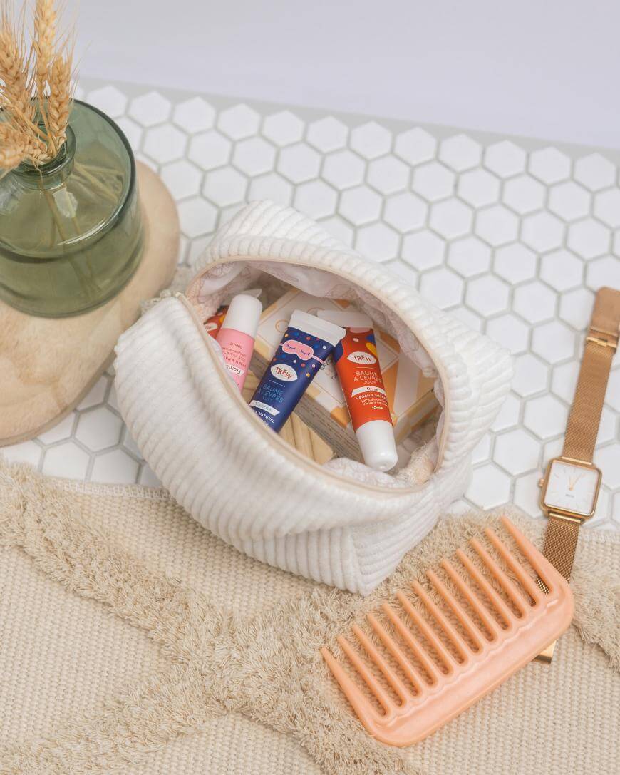 A white corduroy pouch filled with lip balms and small items sits on a textured bathmat, next to an orange comb, a gold wristwatch, and a green glass vase with dried wheat on a white hexagon tile surface.