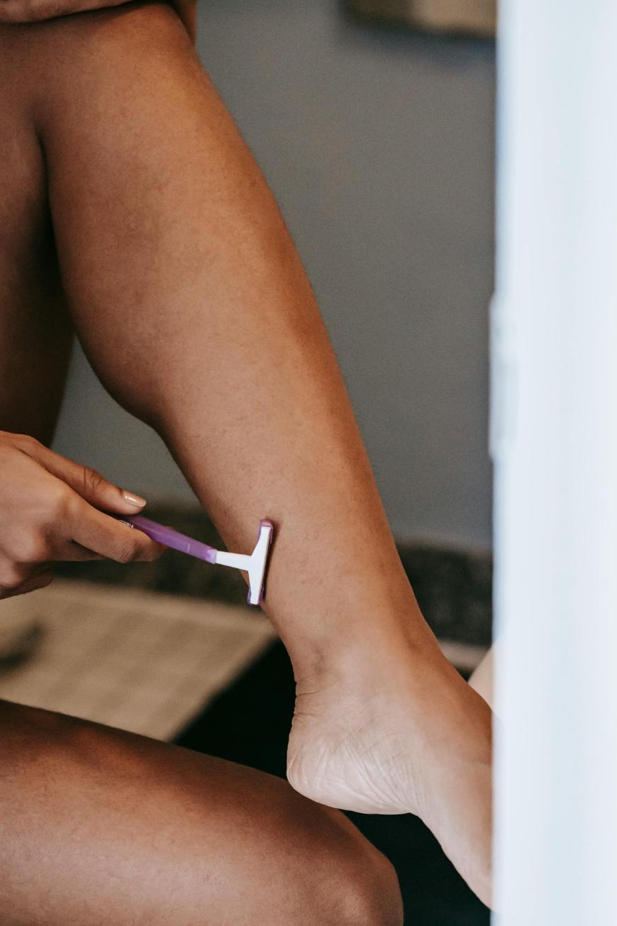 A person is shaving their lower leg with a purple razor. The focus is on their hand holding the razor and their leg, with the rest of the background blurred.