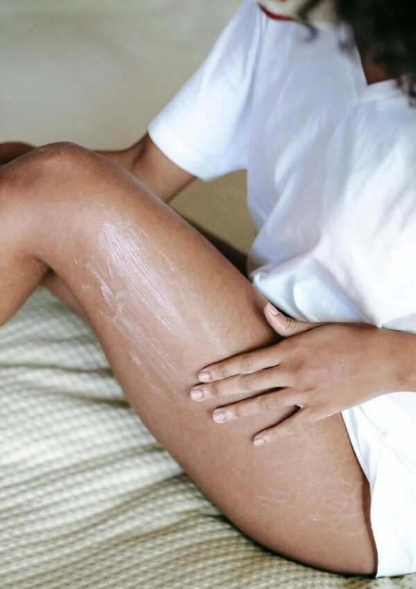A person in a white shirt sits on a bed, applying lotion to their thigh. The focus is on their hands and legs, with visible streaks of lotion on the skin. The background is softly blurred.