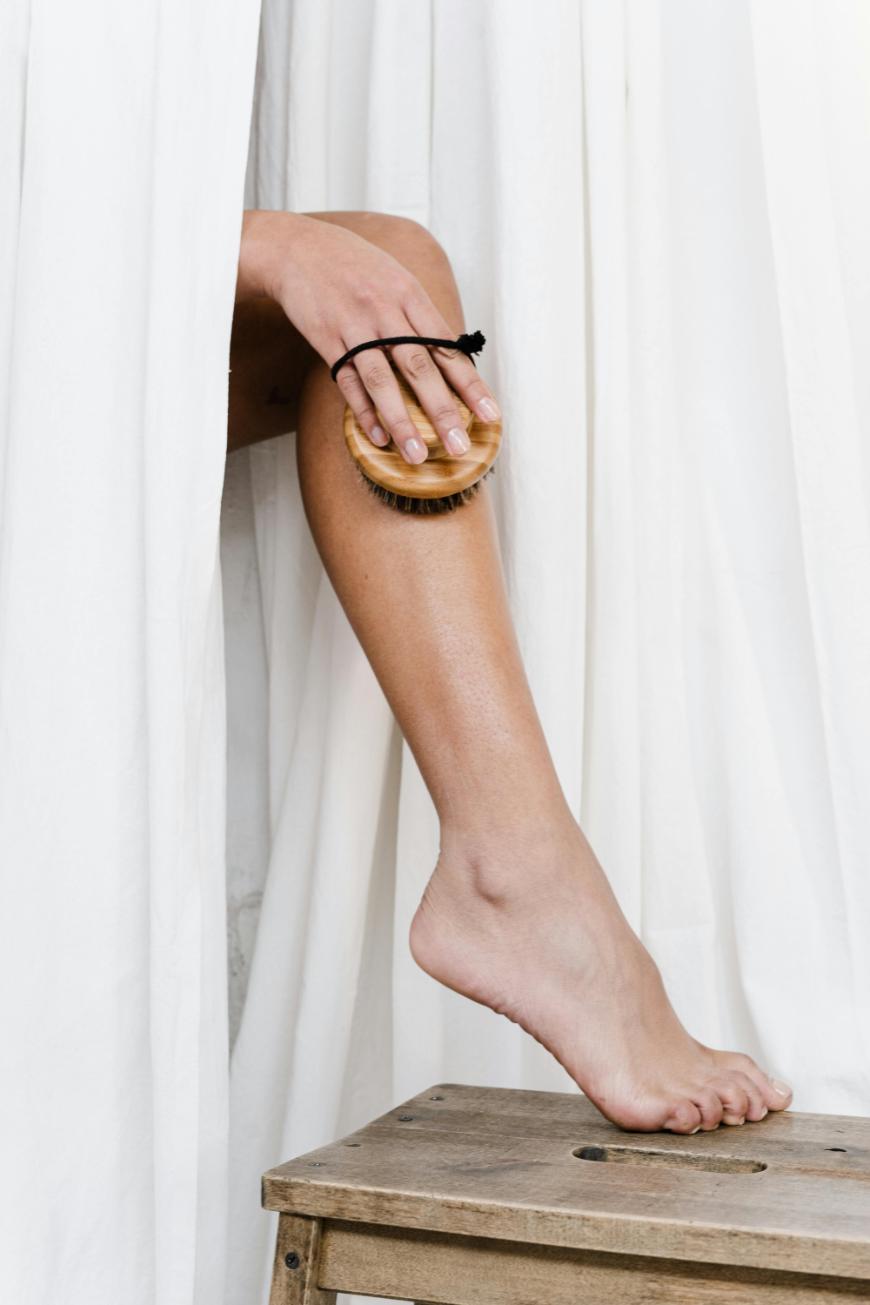 A person standing beside white curtains uses a handheld brush to exfoliate their lower leg, which is resting on a wooden stool.