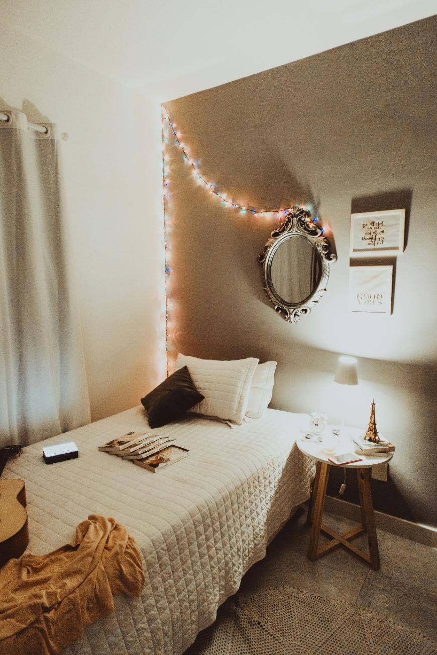 Cozy bedroom with a single bed, neutral bedding, and pillows. A round mirror and two framed artworks hang above the bed. String lights decorate the wall. A bedside table holds a lamp, books, and small decor.
