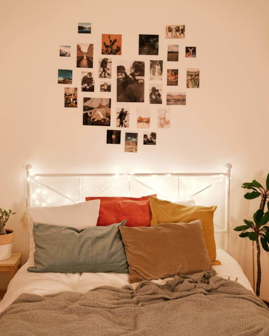 A cozy bed with colorful pillows and a gray blanket sits beneath a headboard decorated with string lights. Above the bed, various photos are arranged on the wall in a loose heart shape. A green potted plant is nearby.