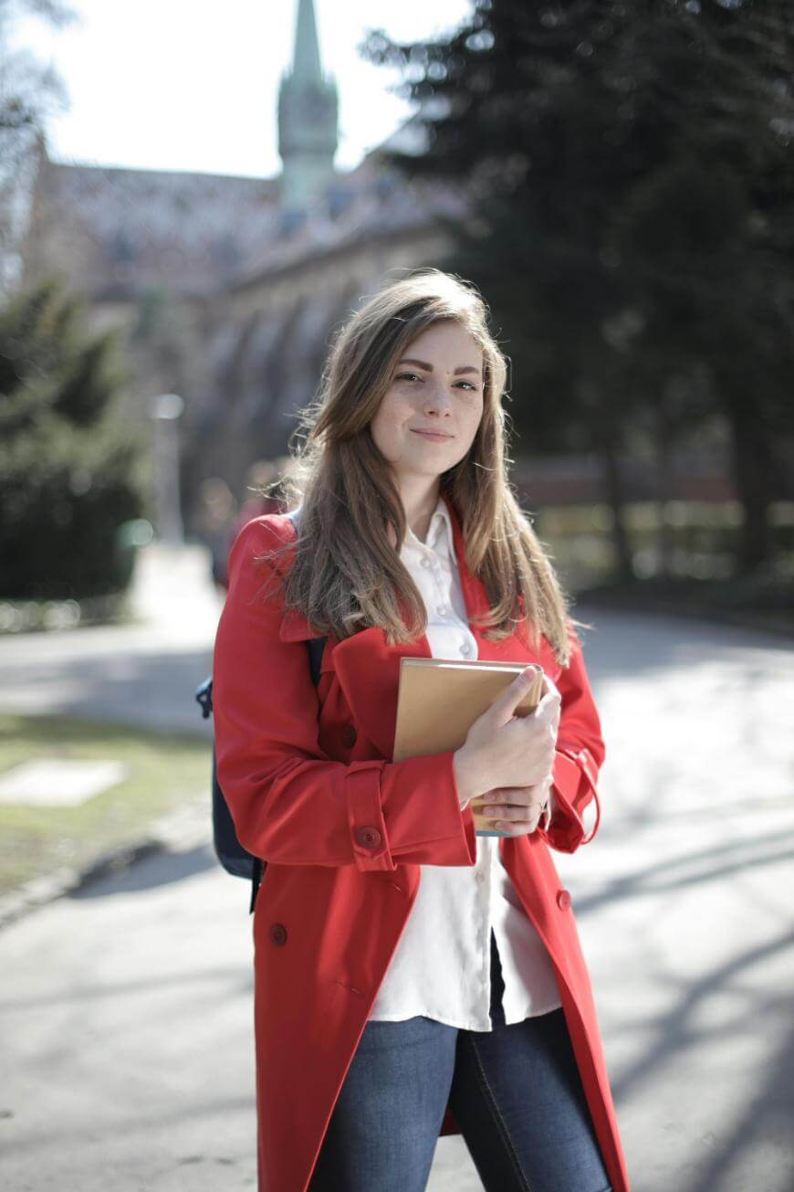 A young woman with long hair, wearing a red coat and white shirt, stands outdoors holding a notebook. She has a backpack on and is smiling slightly. Trees and a building are visible in the background.