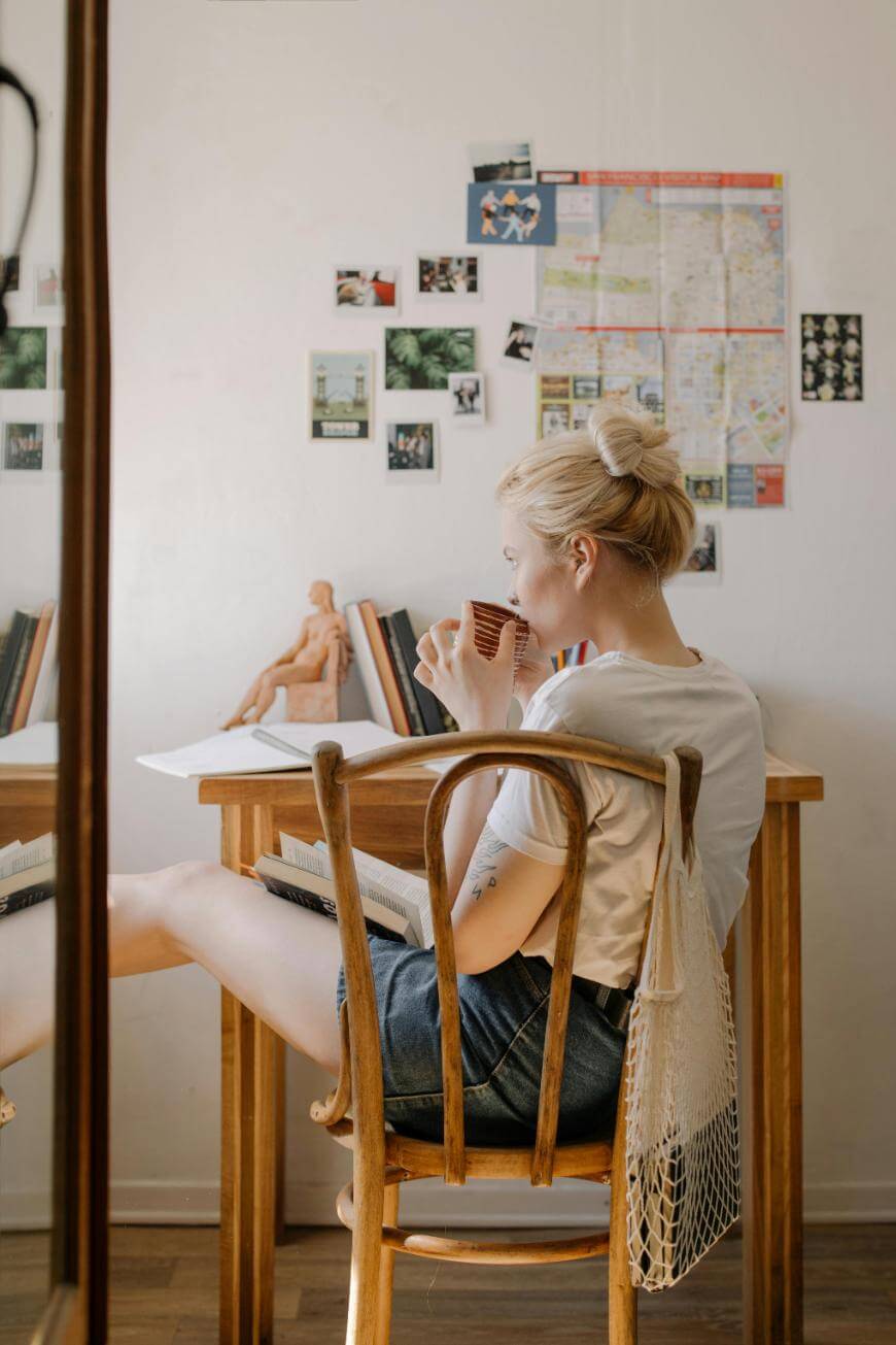 A woman with blonde hair in a bun sits at a wooden desk, sipping from a cup. She has one leg up on the chair, and books, photos, and a map are on the wall behind her. A mesh bag hangs from her shoulder.