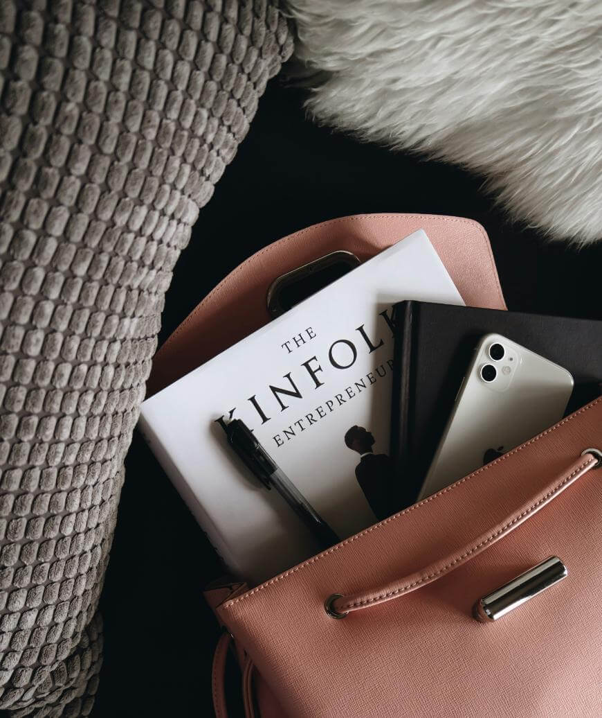 A pink handbag on a textured grey cushion contains a book titled The Kinfolk Entrepreneur, a black notebook, a pen, and a silver iPhone. A fluffy white pillow is also partially visible nearby.
