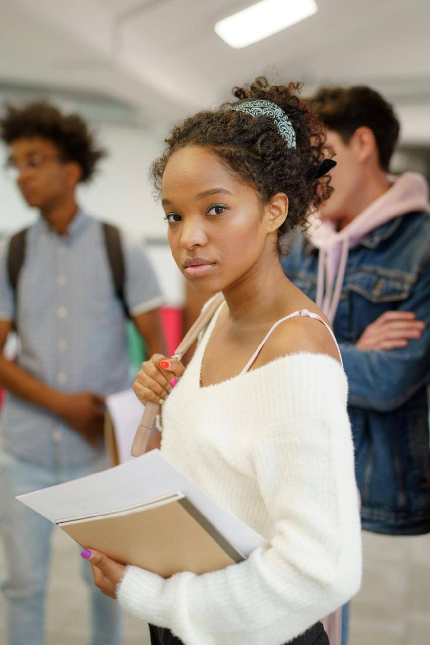 A young woman with curly hair in a white sweater holds notebooks and looks at the camera, standing indoors with two other people blurred in the background.