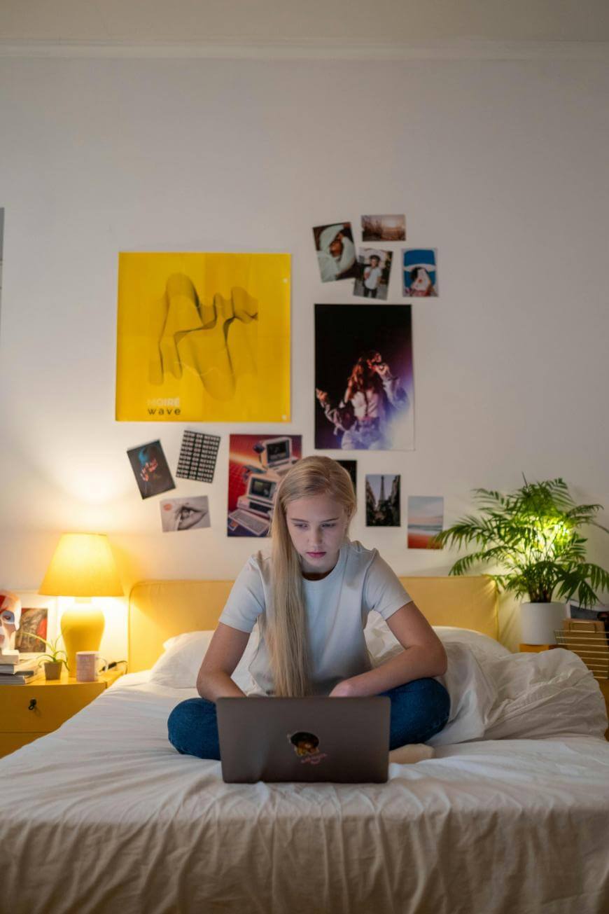A young girl with long blonde hair sits cross-legged on a bed, using a laptop. The room has white walls decorated with posters and photos, a yellow lamp, and a potted plant on a nightstand.