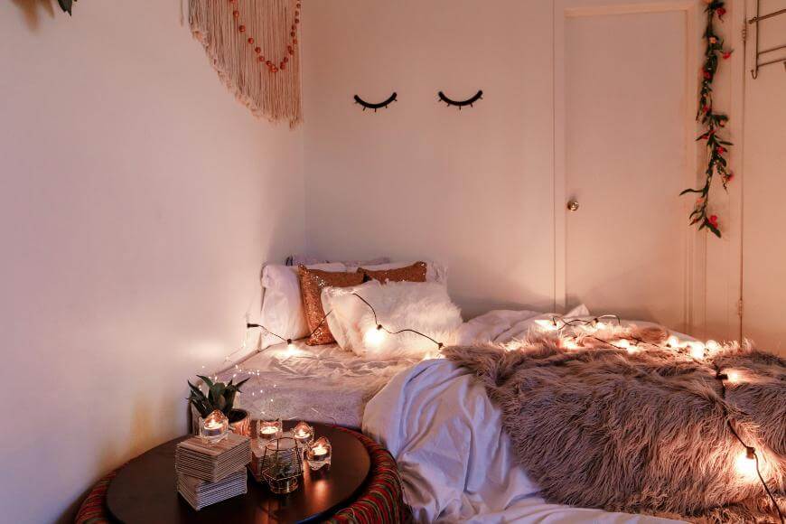A cozy bedroom with a bed covered in soft pillows and a furry blanket, decorated with string lights. There are candles on a round side table, wall art, and eyelash decals above the bed, creating a warm, inviting atmosphere.