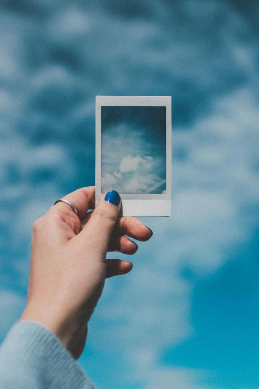 A hand with blue nail polish holds a Polaroid photo of a cloudy sky against a matching real sky background, blending the photo and the scenery together.