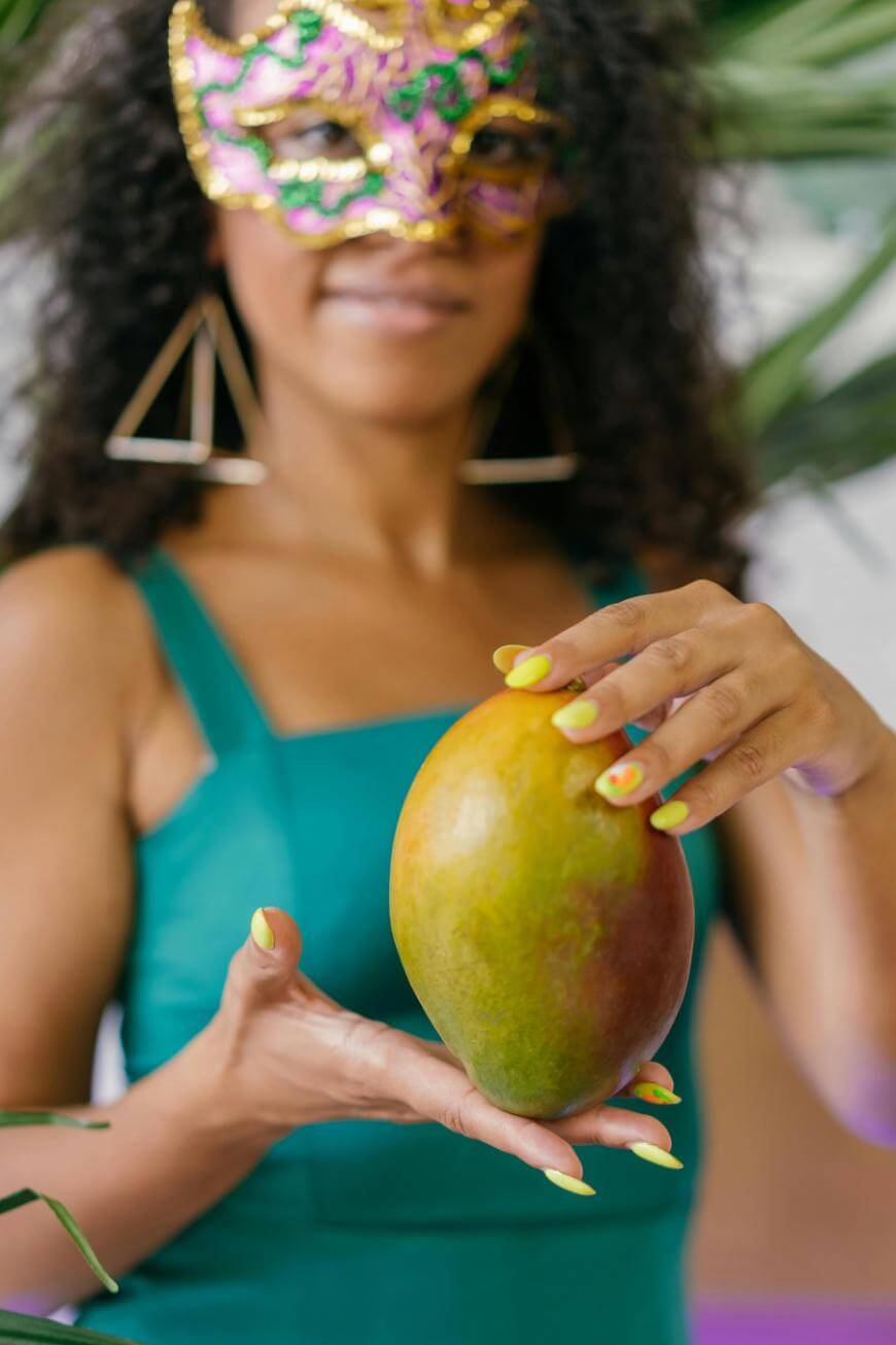 A woman wearing a colorful carnival mask and green dress holds a large mango. Her nails are painted yellow, and she stands among green leaves, partially out of focus.