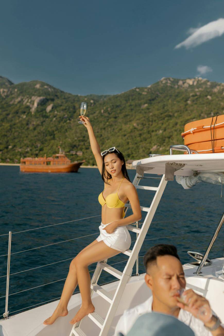 A woman in a yellow bikini top and white shorts poses on a yacht ladder, holding up an empty glass. Behind her, a man sits and drinks. The background features blue water, mountains, and a wooden boat.