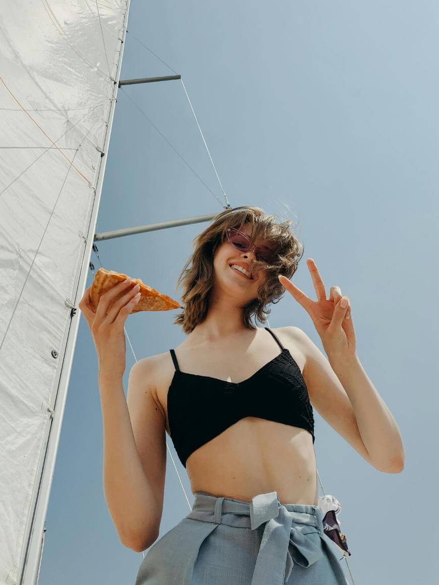 A young woman in a black crop top and blue pants holds a slice of pizza and makes a peace sign, smiling up at the camera. A sail and clear blue sky are visible in the background.