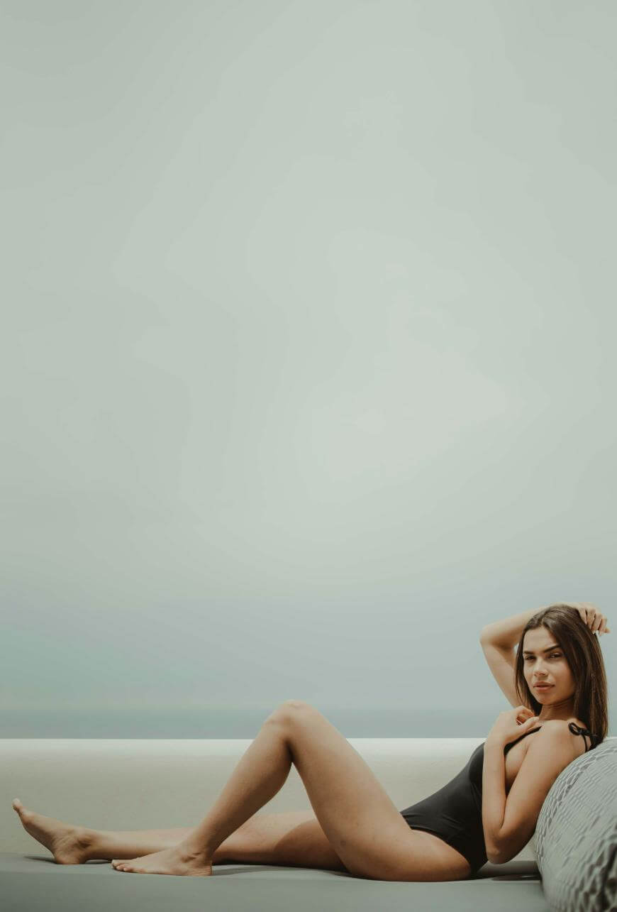 A woman in a black one-piece swimsuit reclines on a light-colored sofa against a minimalist, pale background, looking toward the camera with one hand near her neck.