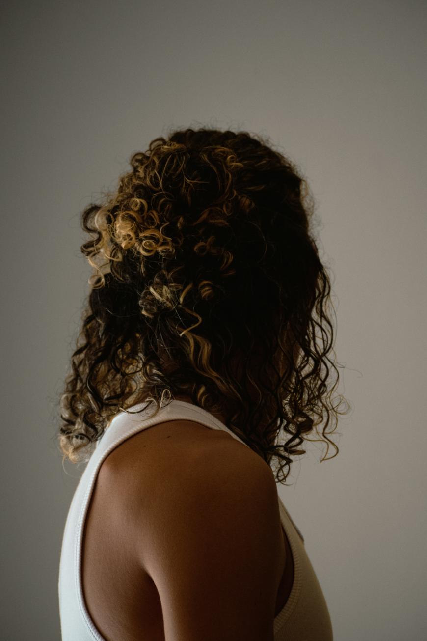 A person with curly, highlighted hair wearing a white sleeveless top stands against a plain background, facing away and slightly to the side, with their face not visible.