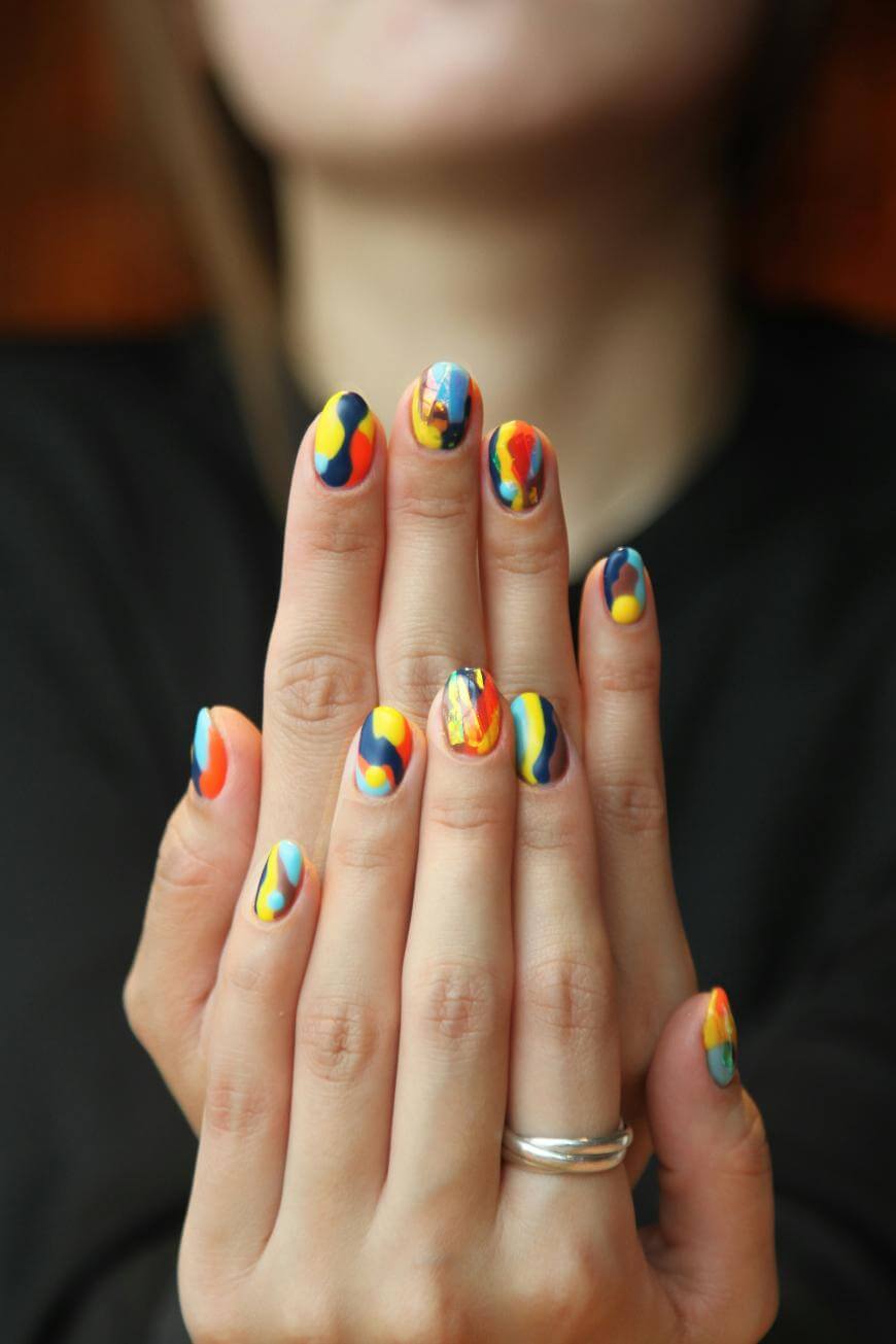 A person with hands pressed together shows brightly colored, abstract nail art featuring blue, yellow, orange, and red swirls. The person wears a silver ring on one finger and has a blurred background.