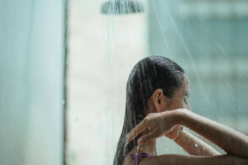 A person with wet hair stands under a running shower, facing away, with one hand raised to their head. Water streams from above and droplets are visible on their skin. The bathroom background is blurred.