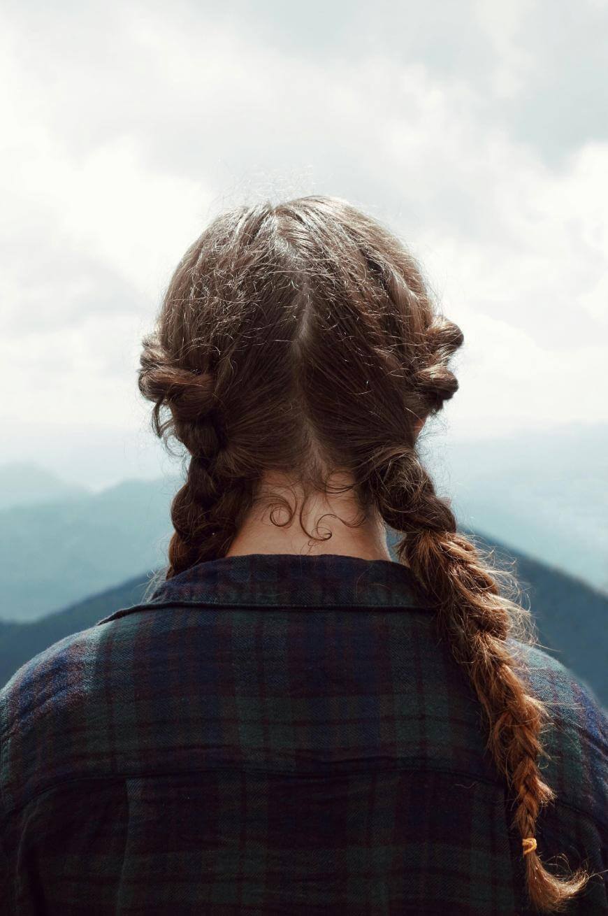 A person with brown hair in two loose braids, wearing a plaid shirt, stands facing away toward a cloudy sky and distant mountains.