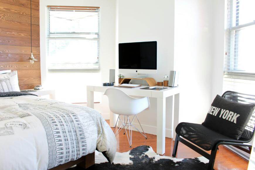 Bright bedroom with a white desk and iMac, white chair, black lounge chair with “New York” pillow, patterned bedding, wooden accent wall, and large windows letting in natural light.