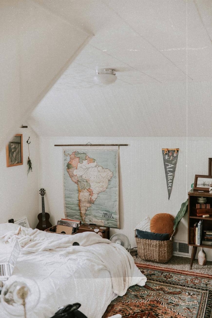 Cozy bedroom with white bedding, a colorful rug, a map of South America on the wall, a guitar, bookshelves, a basket with a cushion, and an Alabama pennant. The room has sloped ceilings and personal decor.