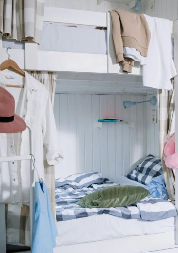 A cozy bedroom with white wooden bunk beds, patterned bedding, and clothes hanging on hangers and rails. A pink hat, green pillow, and folded sweaters add color to the tidy, lived-in space.