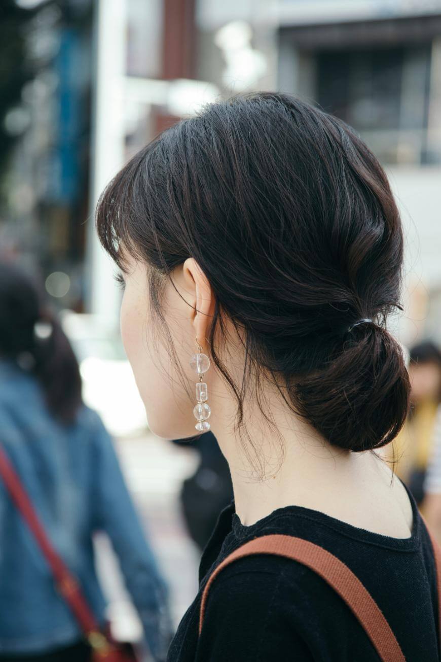 A woman with dark hair styled in a low bun, wearing a black top and dangling clear earrings, stands outdoors with blurred people and buildings in the background. She is seen from behind and slightly to the side.
