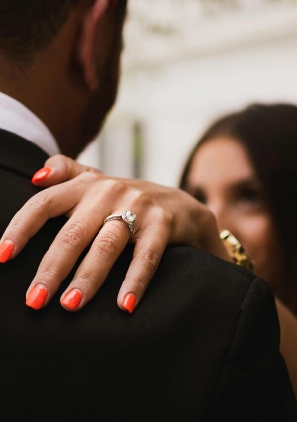 A close-up of a womans hand with bright red-orange nails and an engagement ring resting on a mans shoulder, with both dressed formally and her face softly blurred in the background.