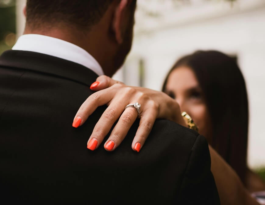 A woman with red nail polish rests her hand, displaying an engagement ring, on a mans shoulder while they embrace, both dressed formally. The background is softly blurred.