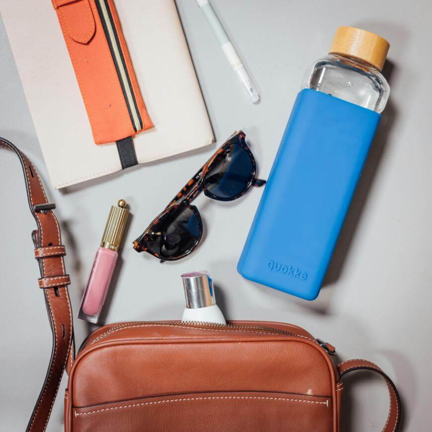 A brown leather bag surrounded by sunglasses, a pink lipstick, a notebook with an orange pen case, a white pen, a blue water bottle with a wooden lid, and a white tube of cream on a light surface.