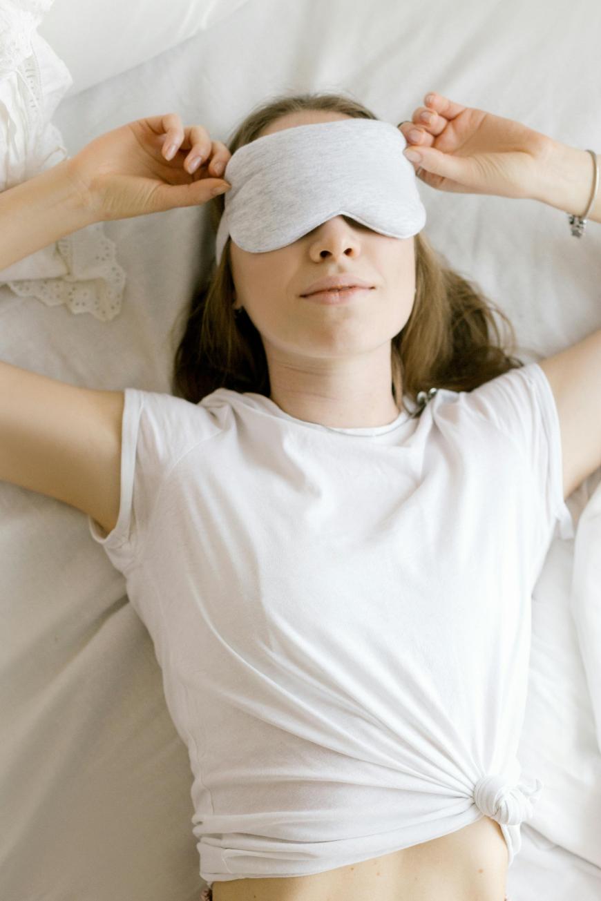 A woman lying on a bed wears a white t-shirt and a light gray sleep mask. She is holding the mask with both hands, appearing relaxed and comfortable, with a soft expression on her face.