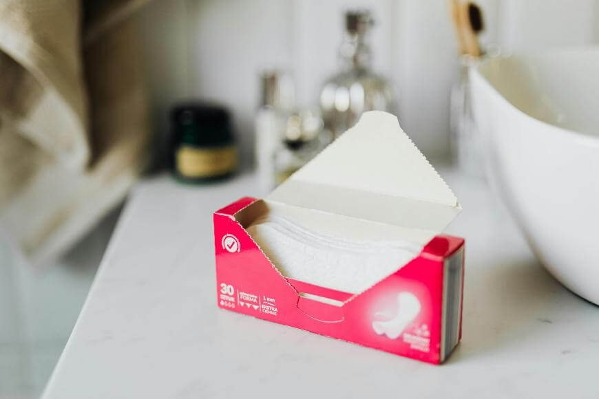 An open pink box of sanitary pads sits on a white bathroom counter near a sink, with toiletries and towels in the blurred background.
