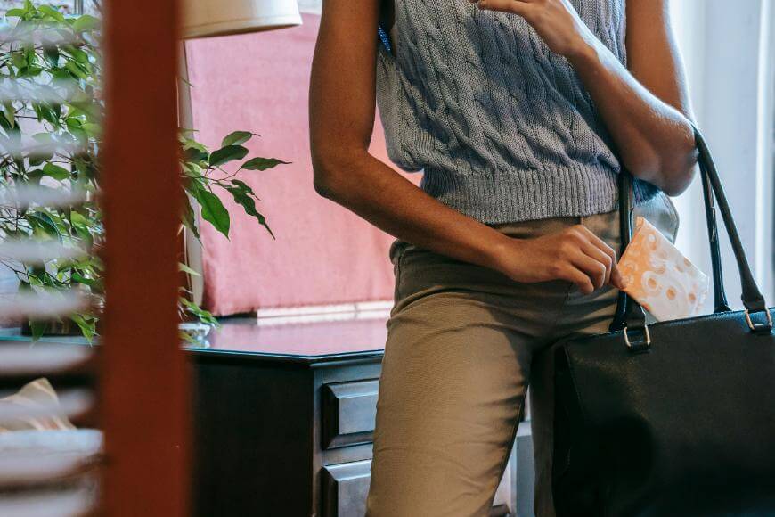 A person wearing a sleeveless knit top and khaki pants holds a black handbag and a face mask while standing indoors near a plant and a window.