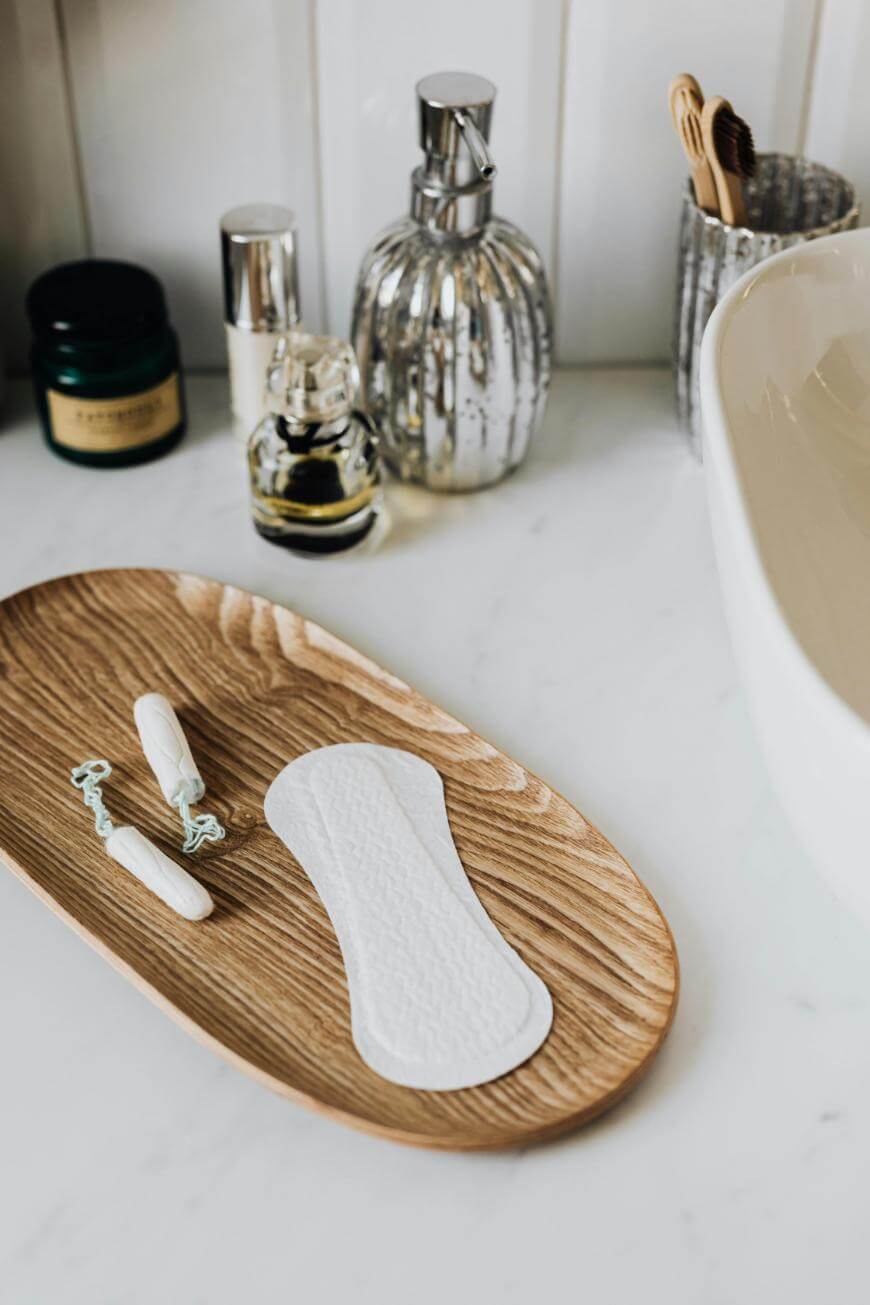 A wooden tray on a bathroom counter holds two tampons and a panty liner. In the background are toiletries, a toothbrush holder with toothbrushes, and a sink.