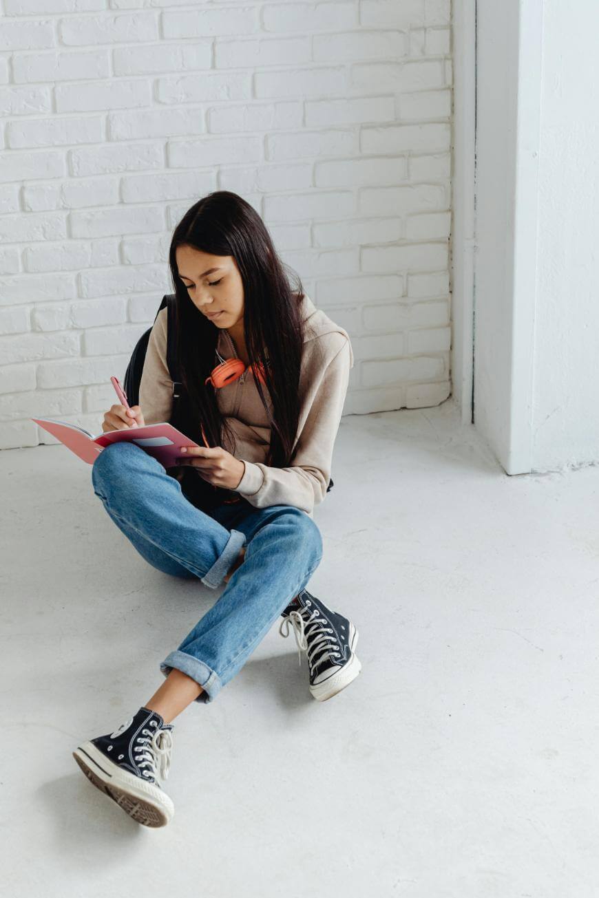 A young woman with long dark hair, wearing a hoodie, jeans, and sneakers, sits on the floor against a white brick wall, writing in a pink notebook. She has a backpack and orange headphones around her neck.