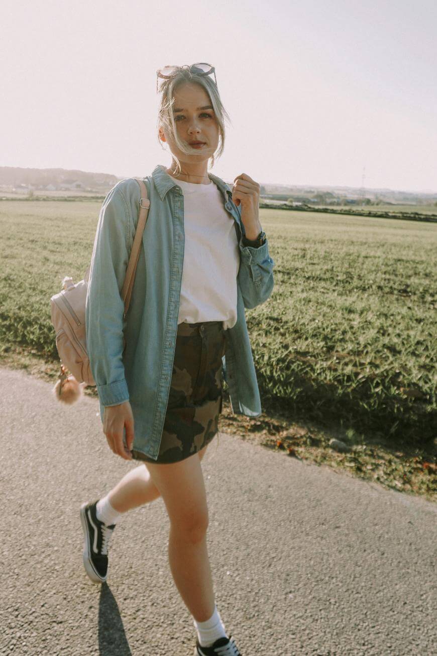 A young woman walks outdoors on a sunny day, wearing a denim shirt over a white t-shirt, a camouflage skirt, sneakers, white socks, and sunglasses on her head, with a backpack over one shoulder. Green fields are in the background.