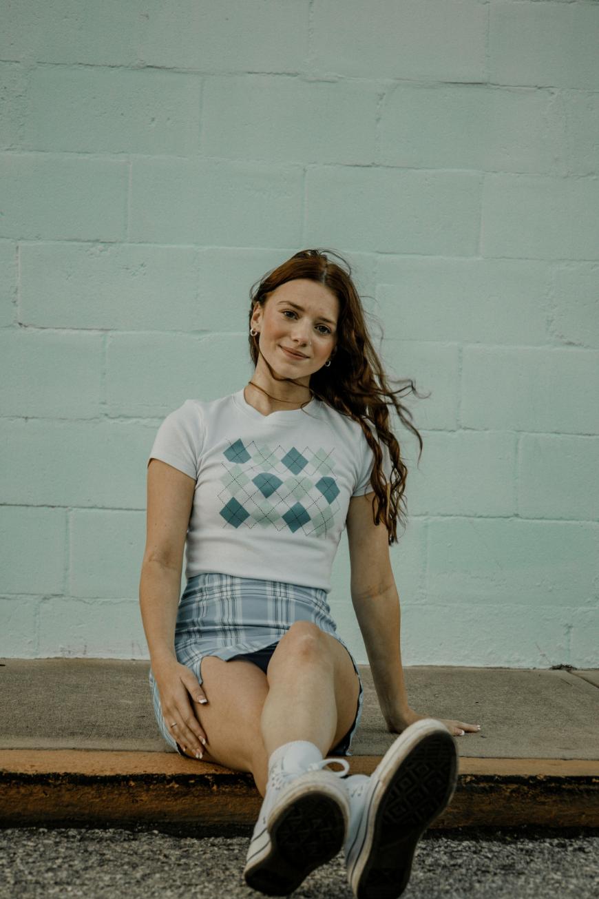 A young woman with long brown hair sits on a sidewalk in front of a pale green brick wall, wearing a white argyle-patterned shirt, a plaid skirt, white socks, and sneakers. She is smiling slightly at the camera.