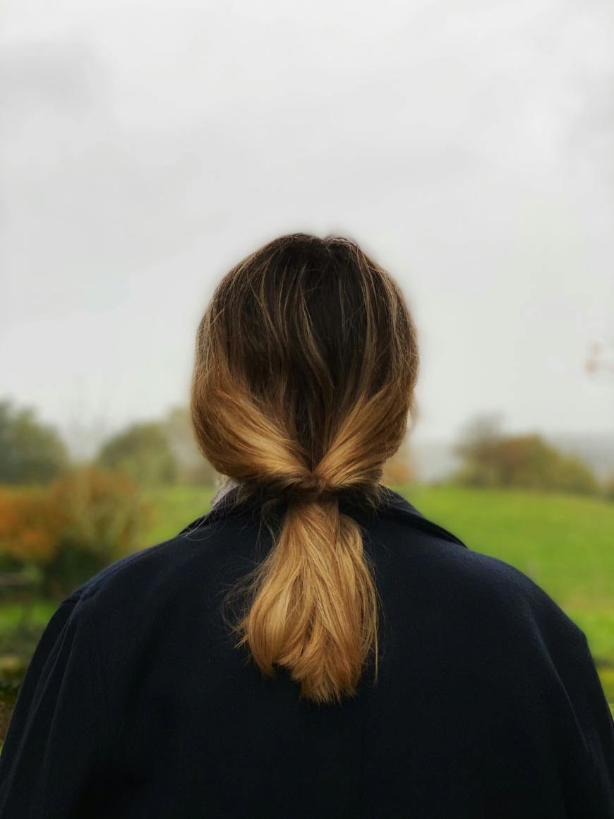 A person with shoulder-length, ombre brown-to-blonde hair is standing outdoors, facing away toward a grassy field on an overcast day, wearing a dark coat.