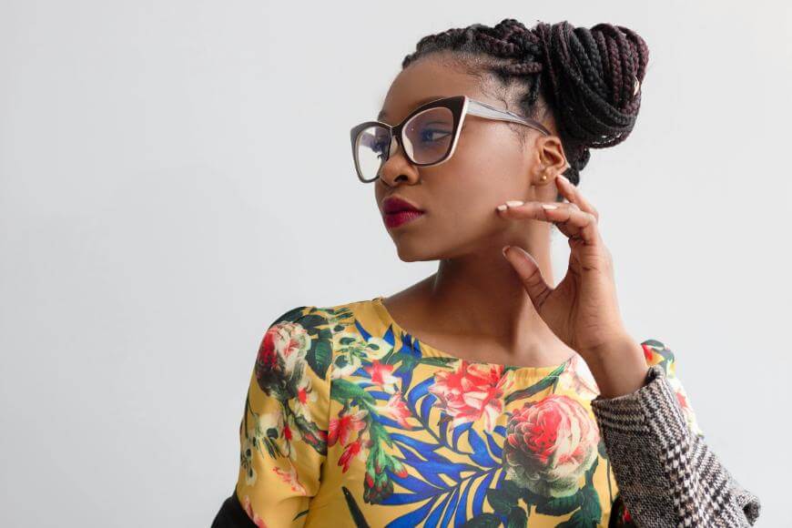 A woman with braided hair in an updo, wearing large square glasses and a yellow floral-patterned top, looks to the side with a confident expression against a plain light background.