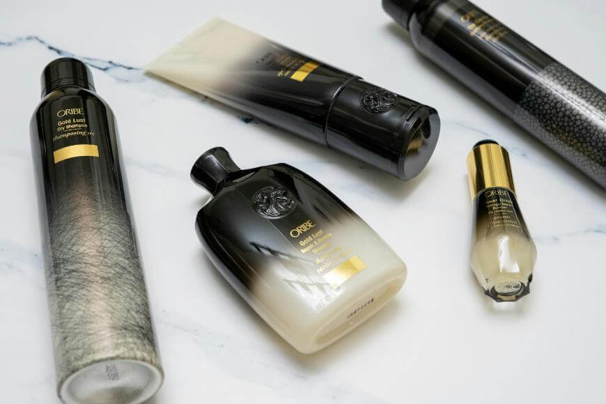 Five Oribe haircare products with black and gold gradient packaging are arranged on a white marble surface. The collection includes bottles, a tube, and a spray can.